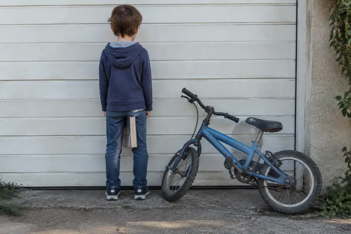 Enfant de dos contre un mur de garage avec un livre entre les jambes, petit vélo usé appuyé à côté